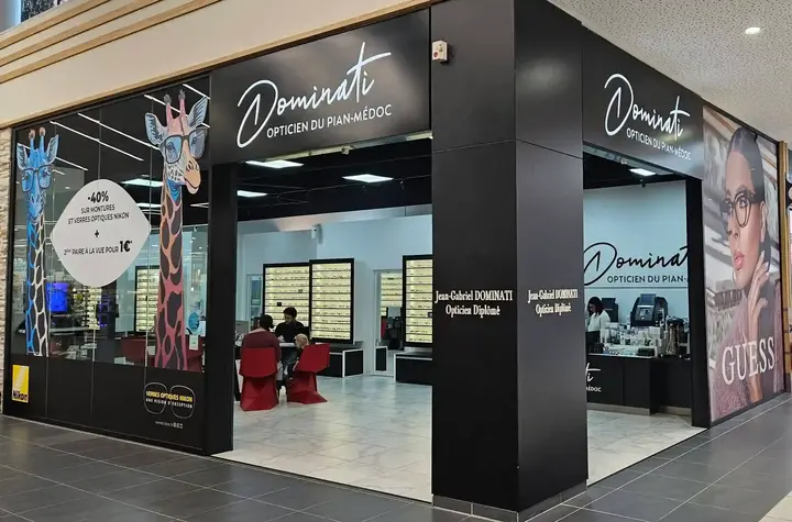 Vue extérieure d'une boutique d'optique 'Dominati Opticien du Pian-Médoc' dans un centre commercial. Le magasin est bien éclairé et affiche une promotion sur les lunettes, avec des présentoirs de lunettes visibles à l'intérieur. Une grande affiche de la marque Guess est également présente.